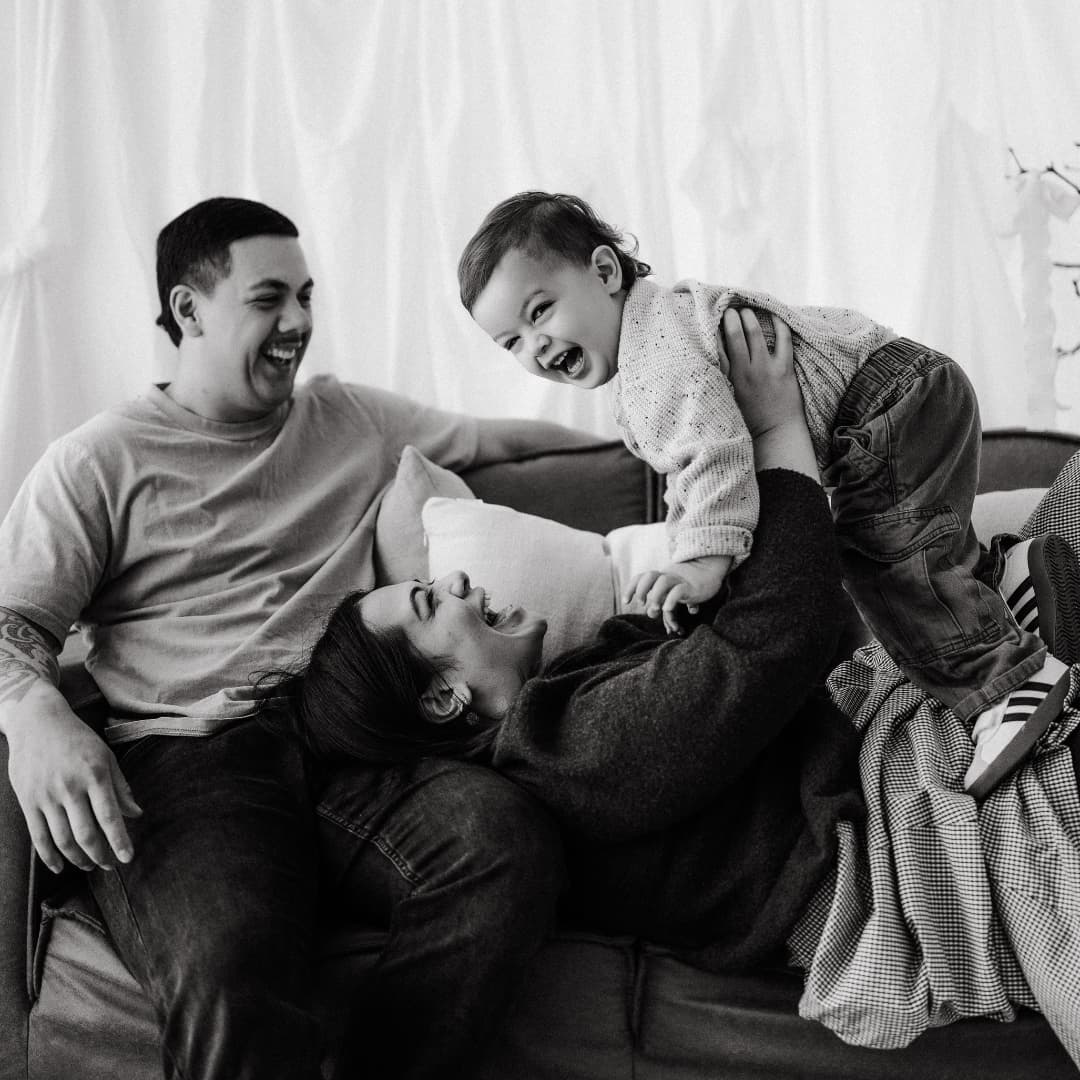 AJ and Tanya, founders of Finer Builds, laughing on the couch with their child — a candid family moment in black and white.